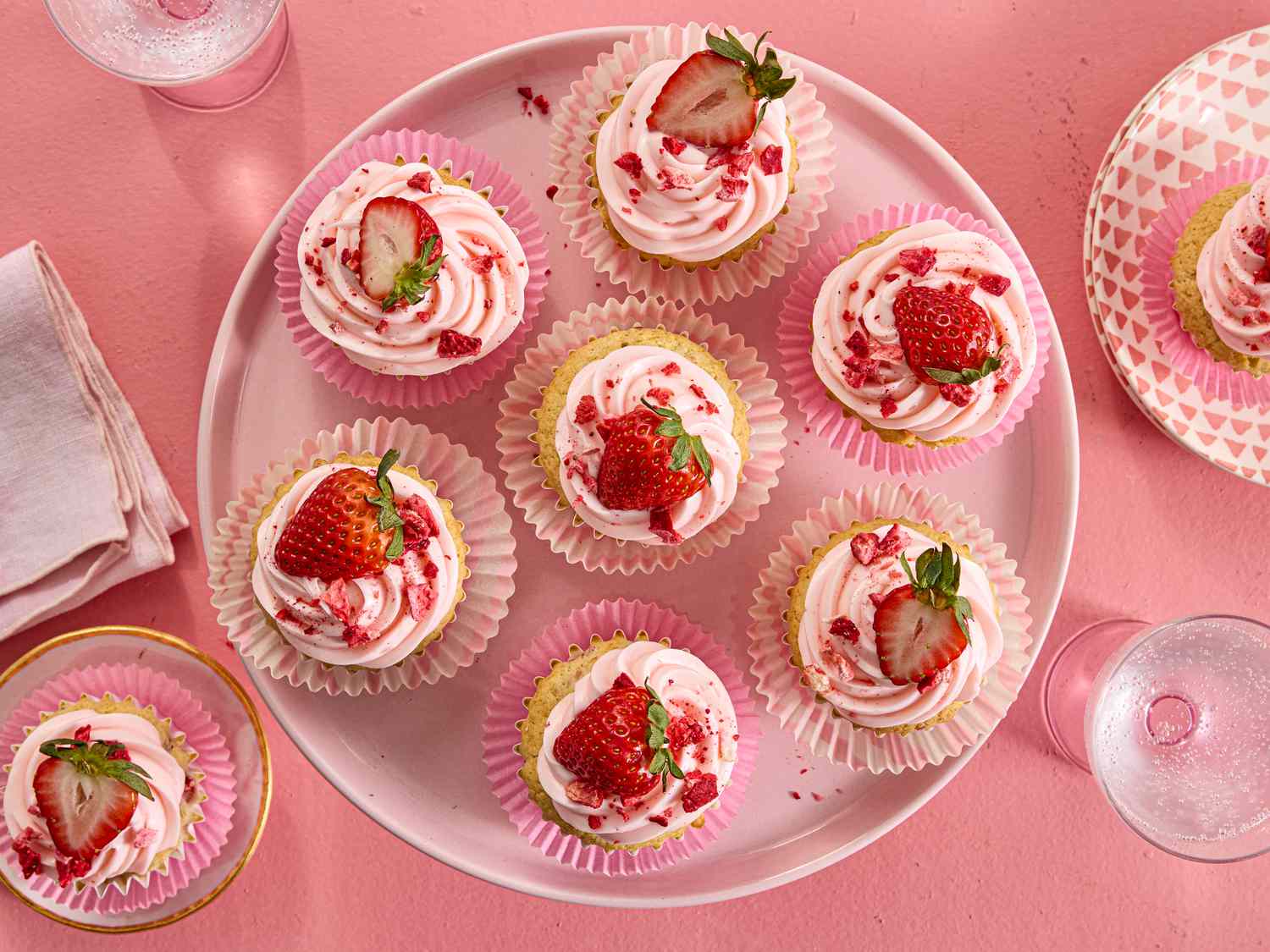 overhead shot of strawberry cupcakes on a pink tray and pink surface. with 2 on trays on the corners