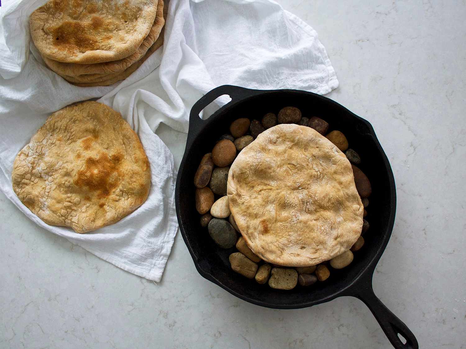 Taboon breads in a cast-iron skillet on river pebbles and spread out across a white linen.