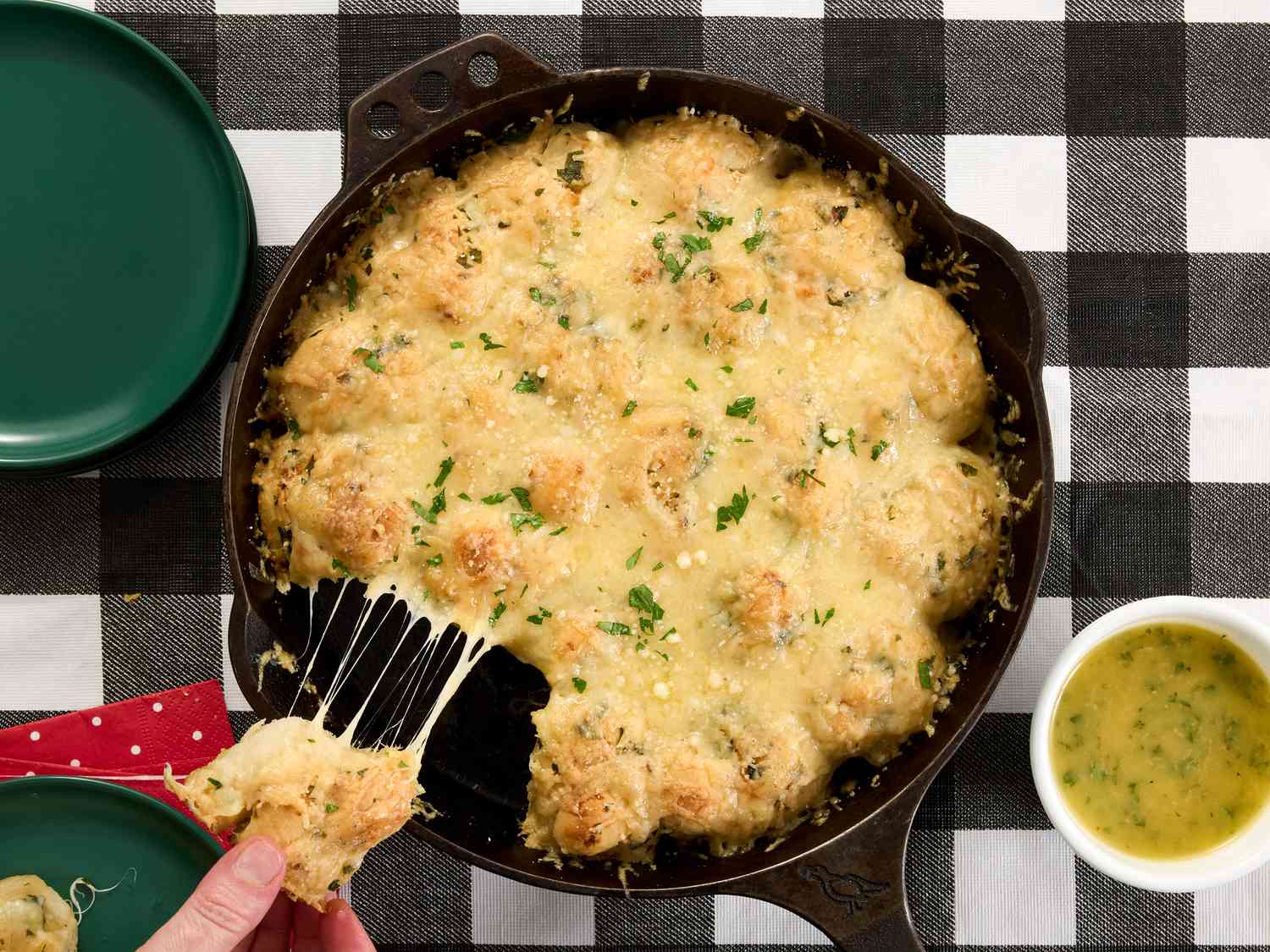 pullinh cheese garlic knot from cast iron pan and small dish of garlic butter on a black and white checkered fabric. 2 green plates and red napkin to the sides. 