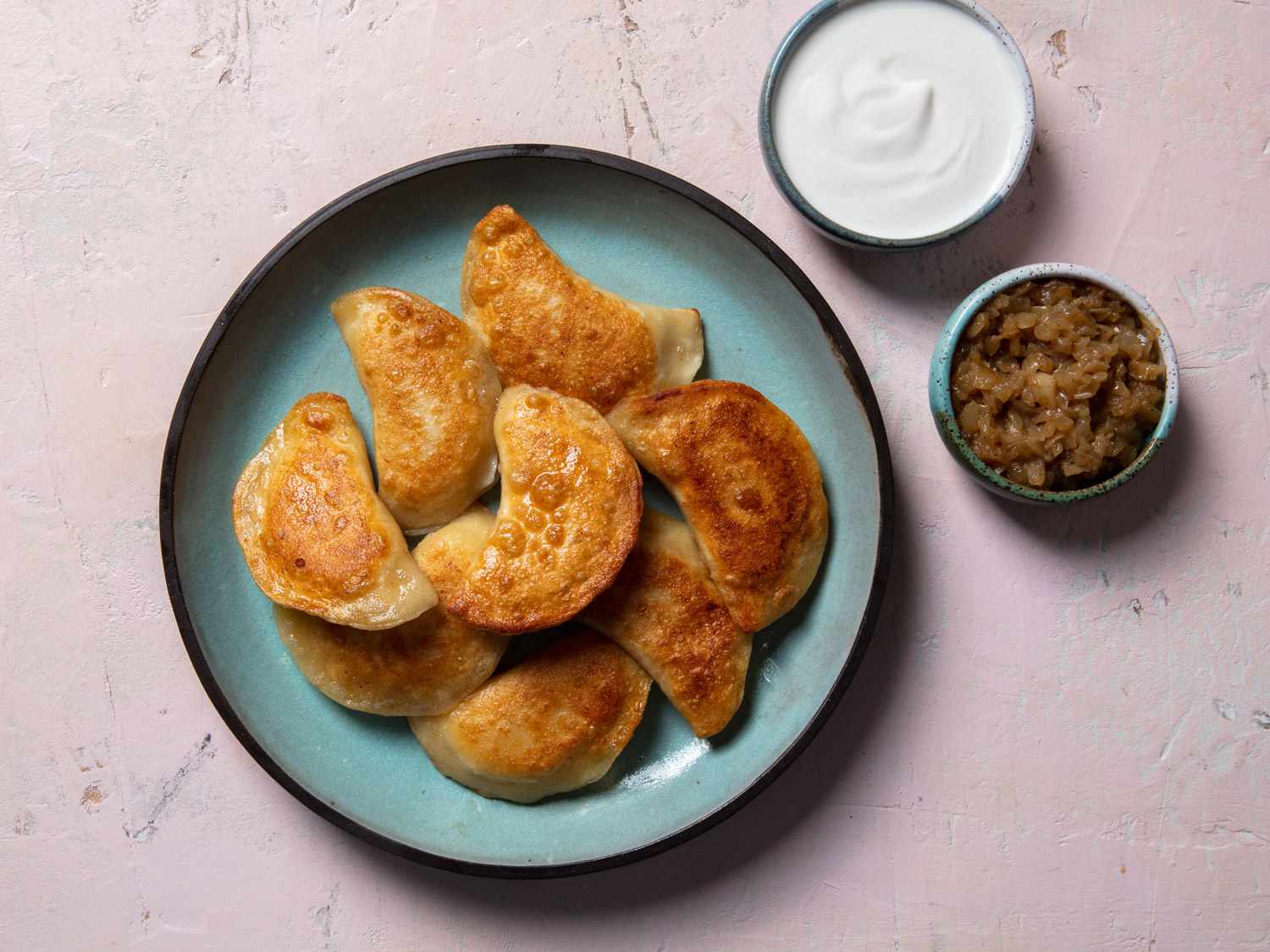 Serving plate of pan-fried pierogi with ramekins of sour cream and cooked onion on the side.