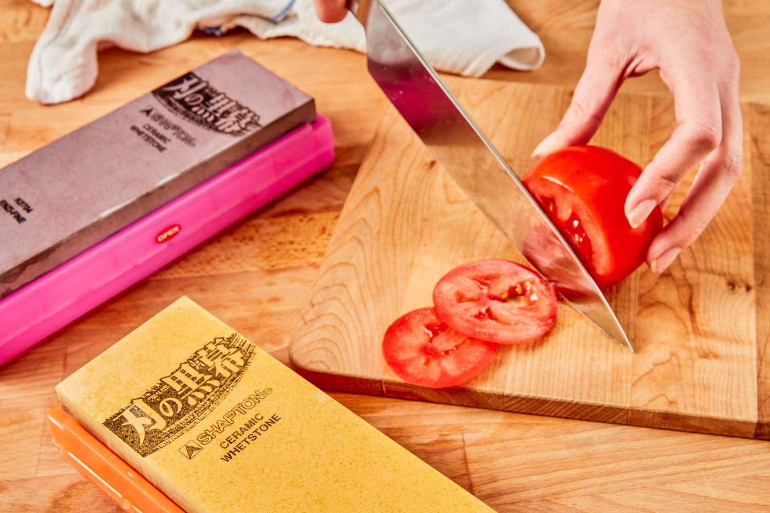 A person slices a tomato with a chef's knife. Two knife sharpeners are also in the frame.