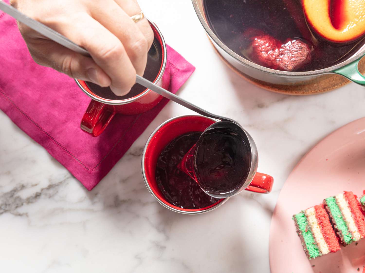 Ladle pouring mulled wine into red mugs, maroon napkin, and pink plate of christmas colored cookies