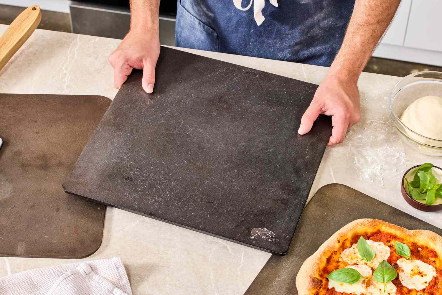 A person placing a pizza steel onto a kitchen countertop. A cooked pizza is on one of the pizza steels.