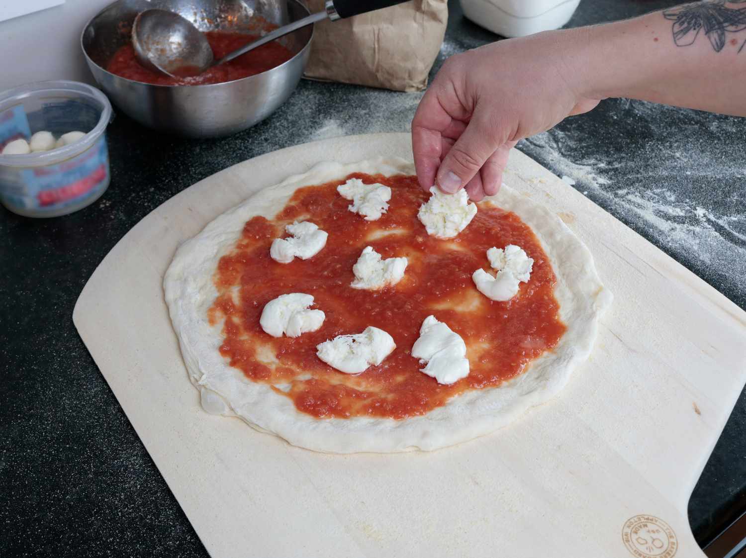 A hand laying mozzarella on a pizza atop a wooden pizza peel