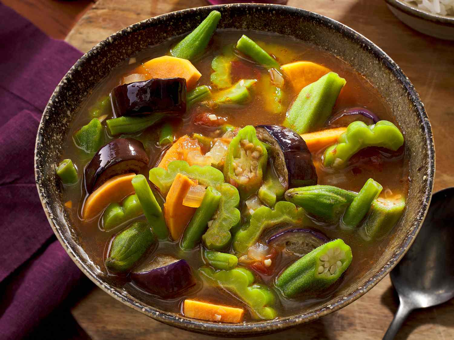 Bowl of pinakbet stew on a wooden surface, with a purple napkin to the left and a spoon to the right 