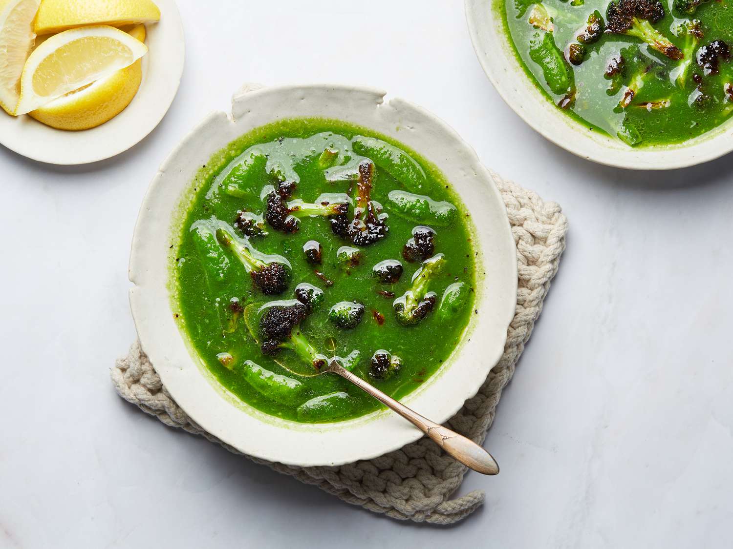 Green Broccoli, leek and snap pea soup from above on a small woven hot pad, with lemons and a second bowl to the side 