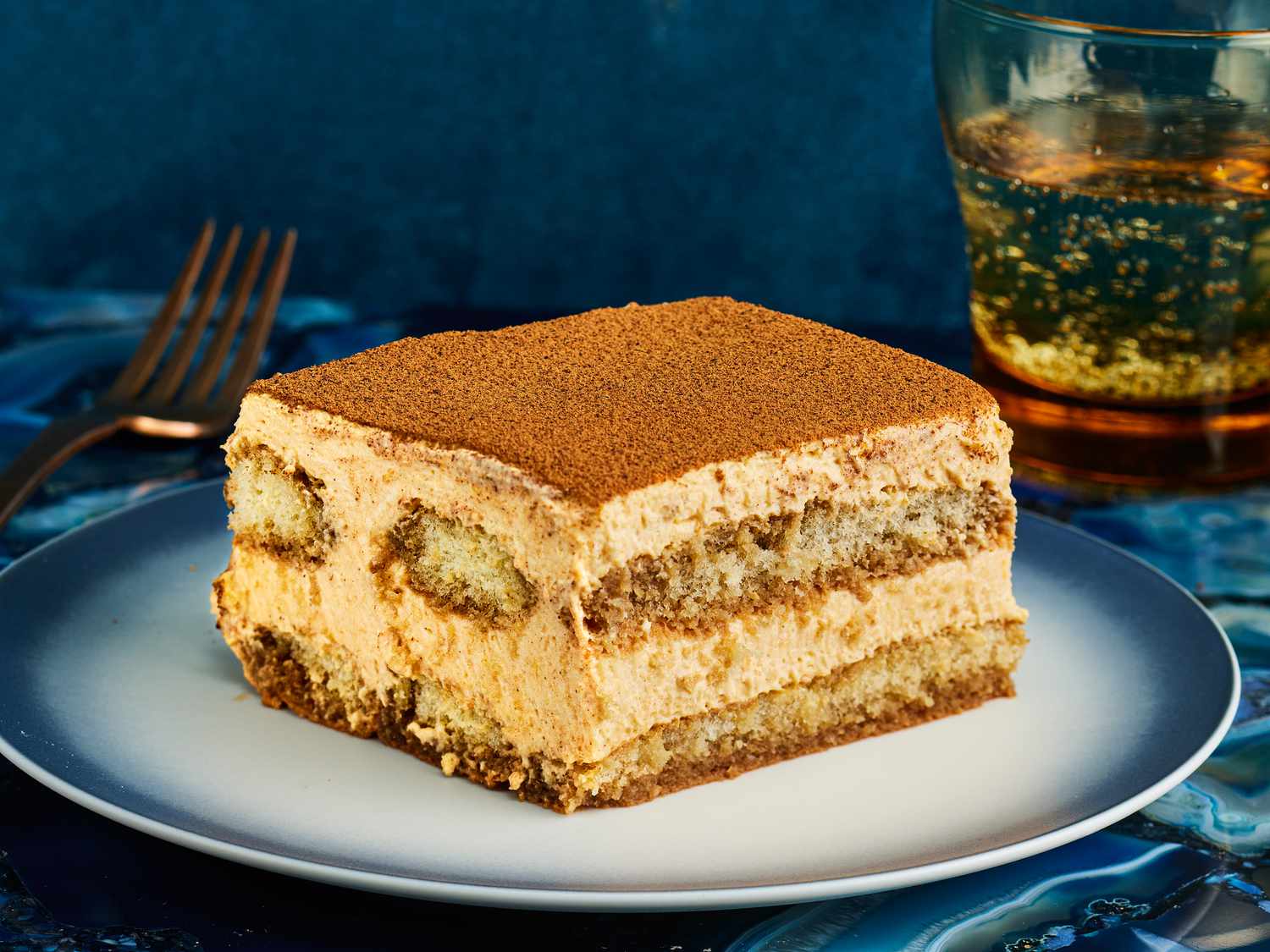 Slice of PSL Tiramisu on a stylish blue and white plate, with a blue tablecloth, sparkling water, and a gold fork