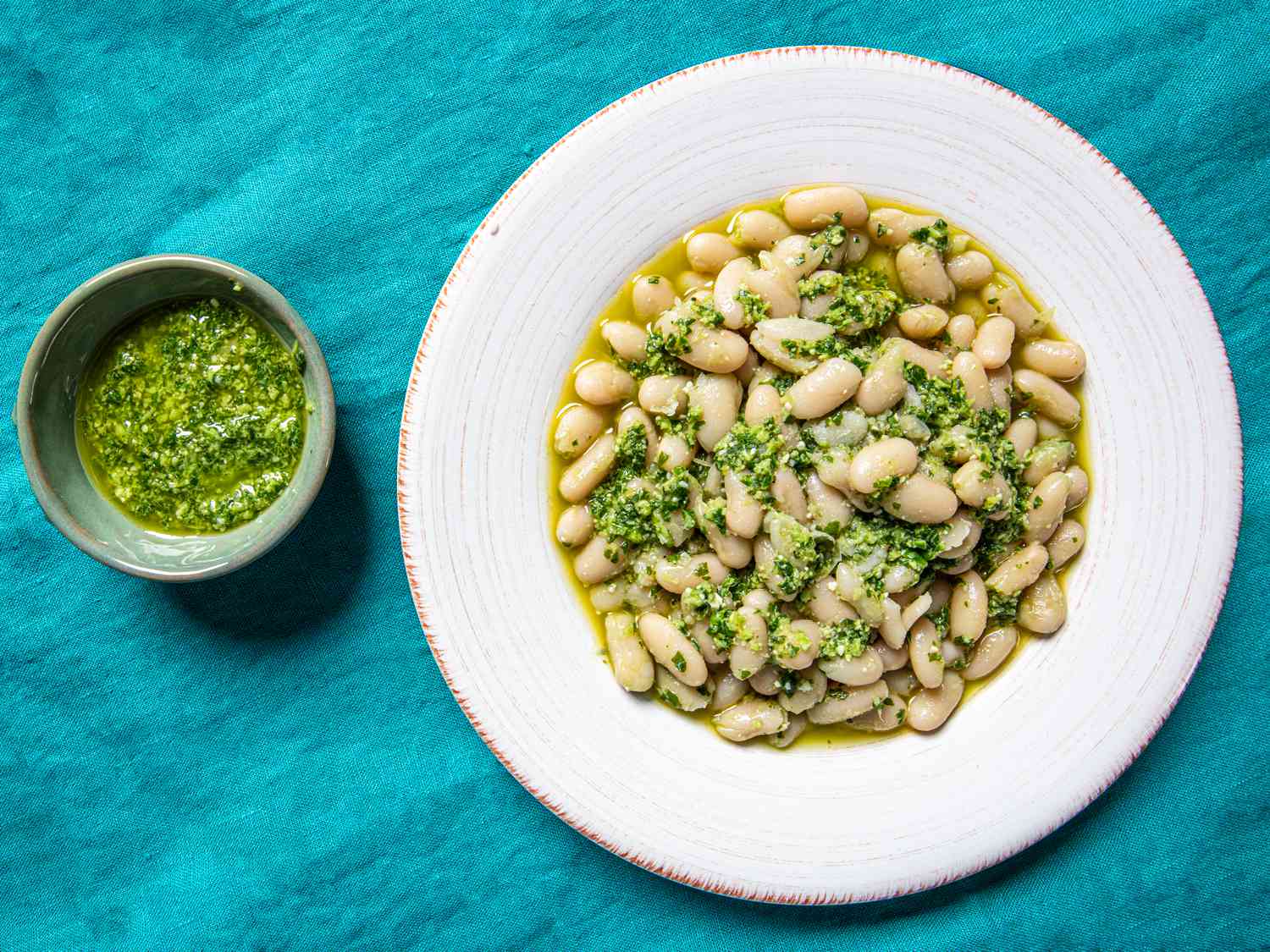 Overhead view of garlic scape pesto in a small bowl next to a wide rimmed, shallow bowl of cooked white beans and pesto