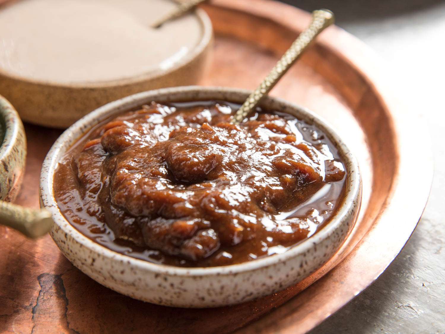 A wide, shallow cream-colored ceramic bowl with brown speckles of glaze on it, holding chunky tamarind chutney and a metal spoon. The bowl is placed on a copper tray and in the top left corner of the image is another similar bowl holding a different sauce.