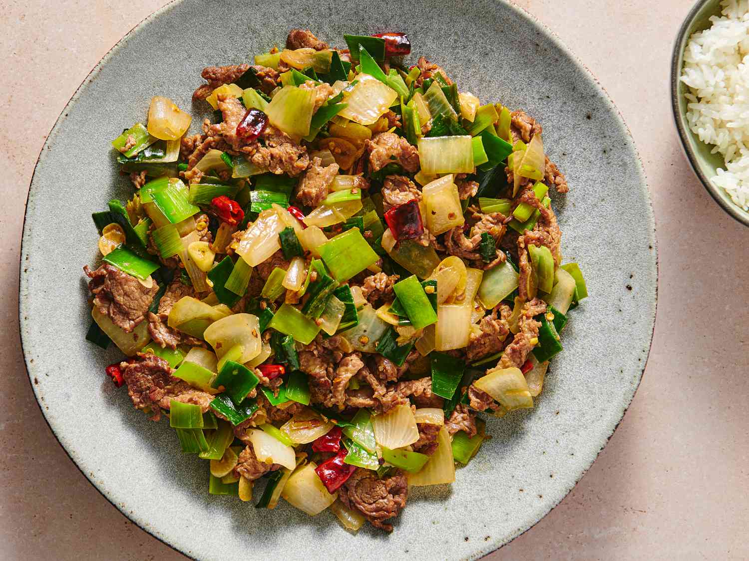 A speckled ceramic plate holding a serving of spicy stir-fried beef with leeks and onions. There is a small bowl of white rice in the top right corner of the image.