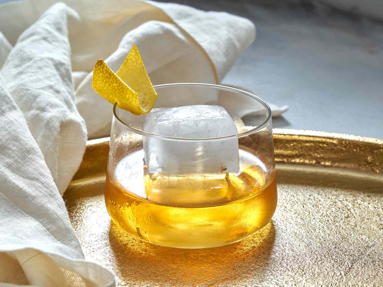 Rusty nail cocktail, with a large ice cube and an orange peel garnish on a gilded tray.