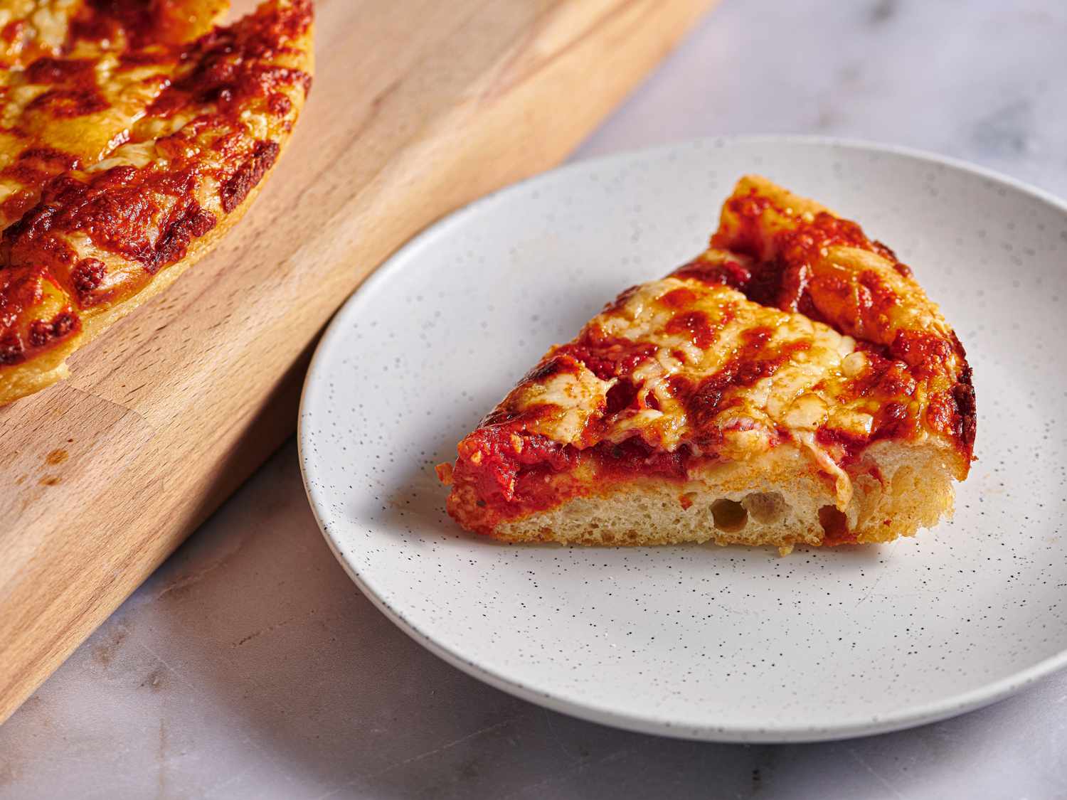 A slice of New England Greek-Style pizza on a speckled ceramic plate. There is a cutting board holding more pizza on the left hand side of the image