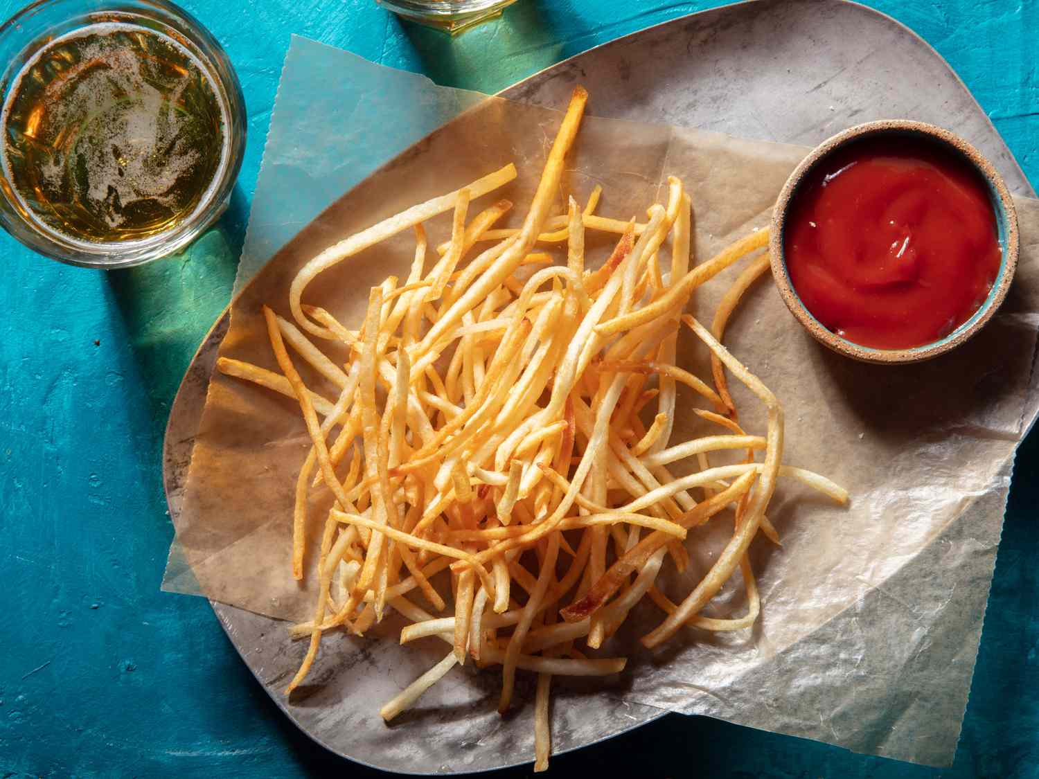 Overhead view of shoestring fries on wax paper on a platter with a ramekin of ketchup and a glass of beer alongside