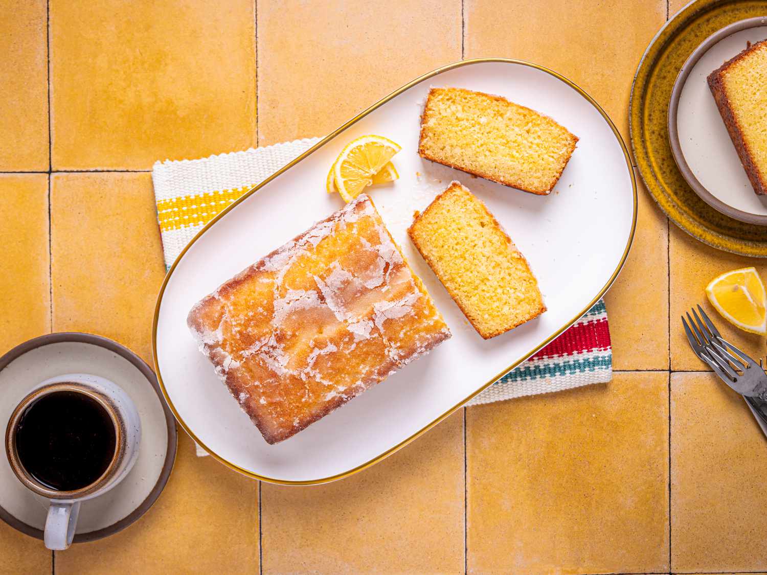 Platter with sliced lemon cake on yellow tile. On the side is a slice of cake, forks, and coffee mug