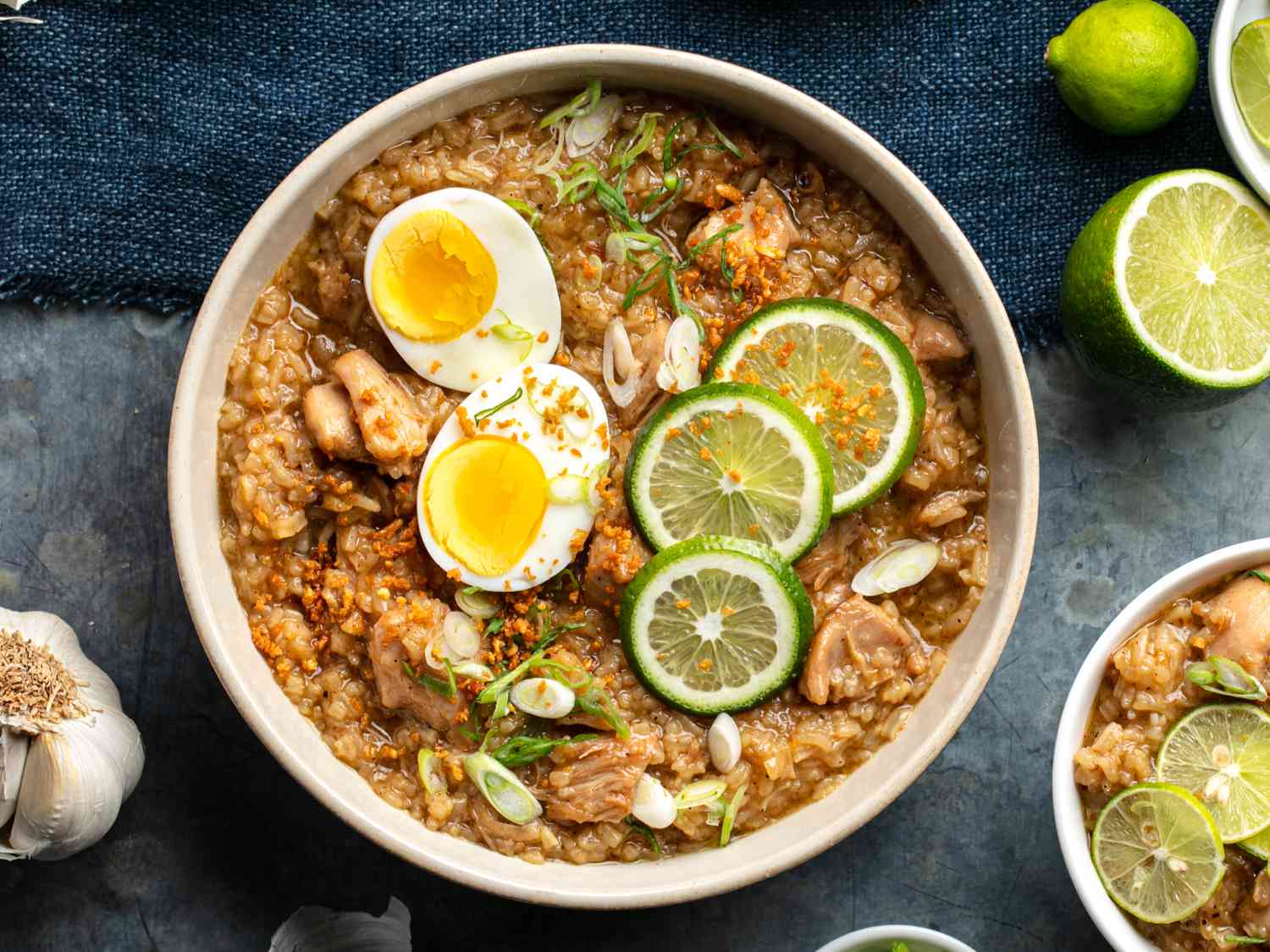 An overhead photo arroz caldo (Filipino chicken and rice soup), topped with a split boiled egg, lime wheels, and sliced scallions. A bulb of garlic and a halved lime is next to the bowl.