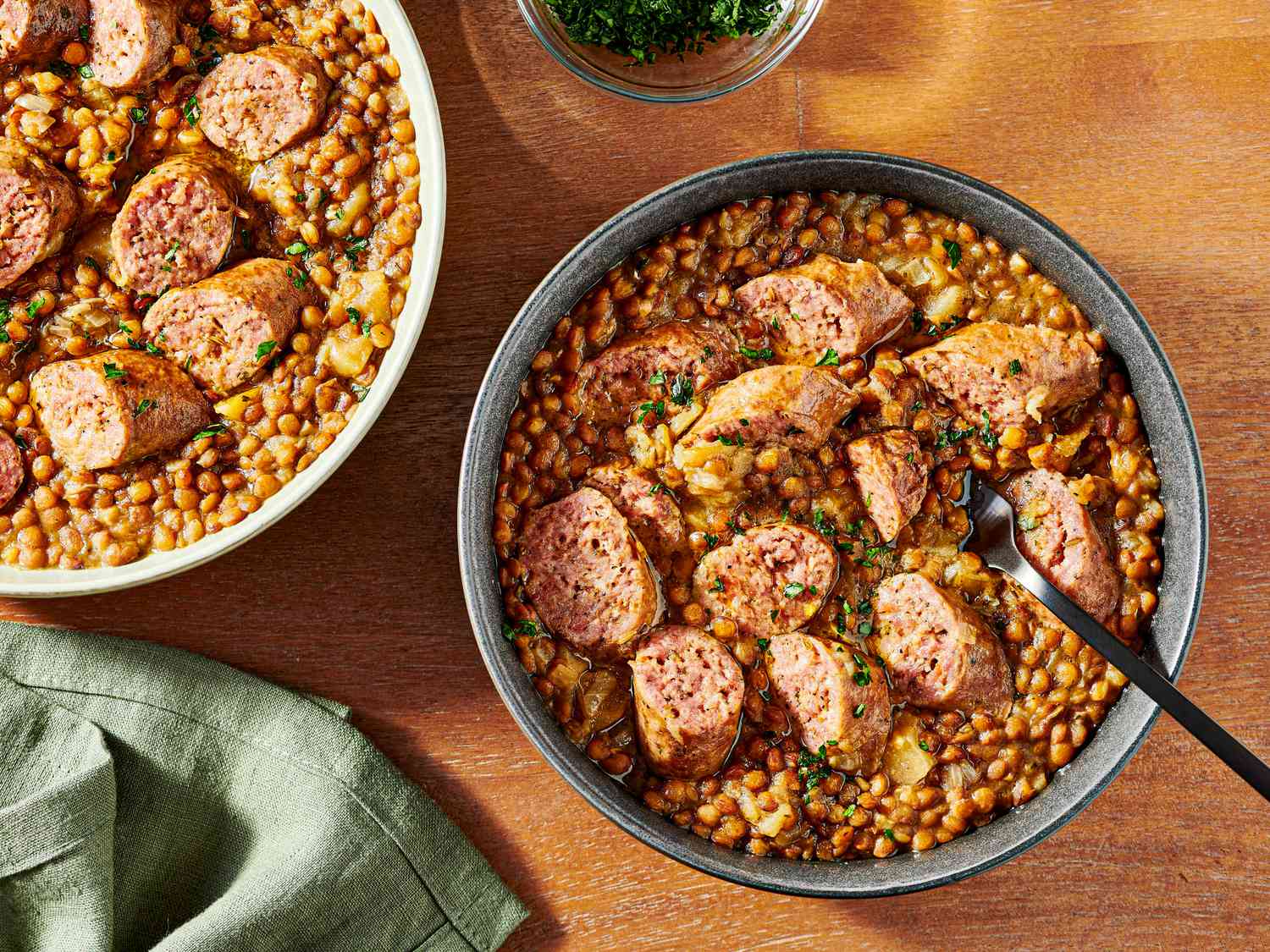 2 bowls of pressure cooked lentils and sausage on a wooden tabletop, with a soft turquoise napkin, and small bowl of parsley for garnish to the sides. 