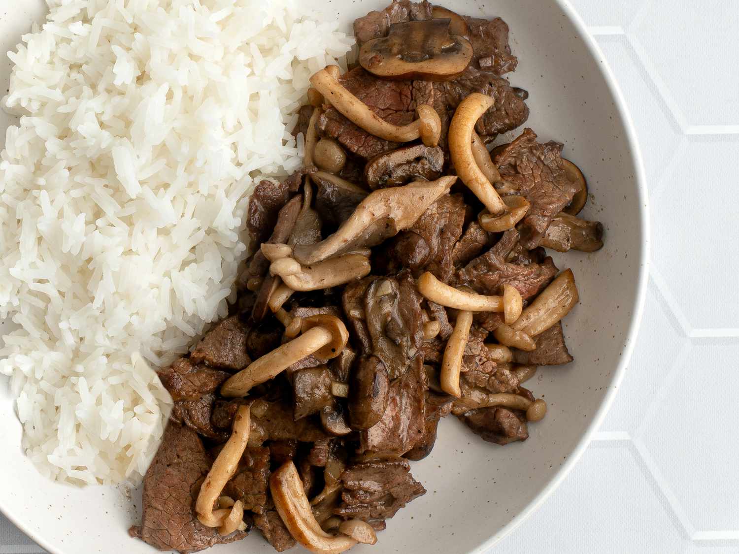 A closeup shot of stir-fried beef with mushrooms and butter. The beef and mushrooms are plated in a round white bowl with white rice on a white tile background, and the shot shows how the mushrooms have been seared, while the feed is tender and in a somewhat glossy sauce.