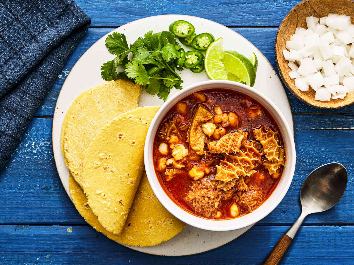 Overhead view of menudo rojo in a bowl on a plate with three taco shells, cilantro, jalapeno and lime wedges. The plate is on a blue table, next to a bowl of diced onion and a spoon. 