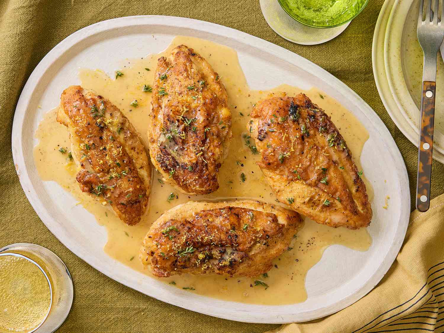 Lemon Pepper chicken on a white platter on a mustard colored textile, with yellow napkin, plates and drinks surrounding it