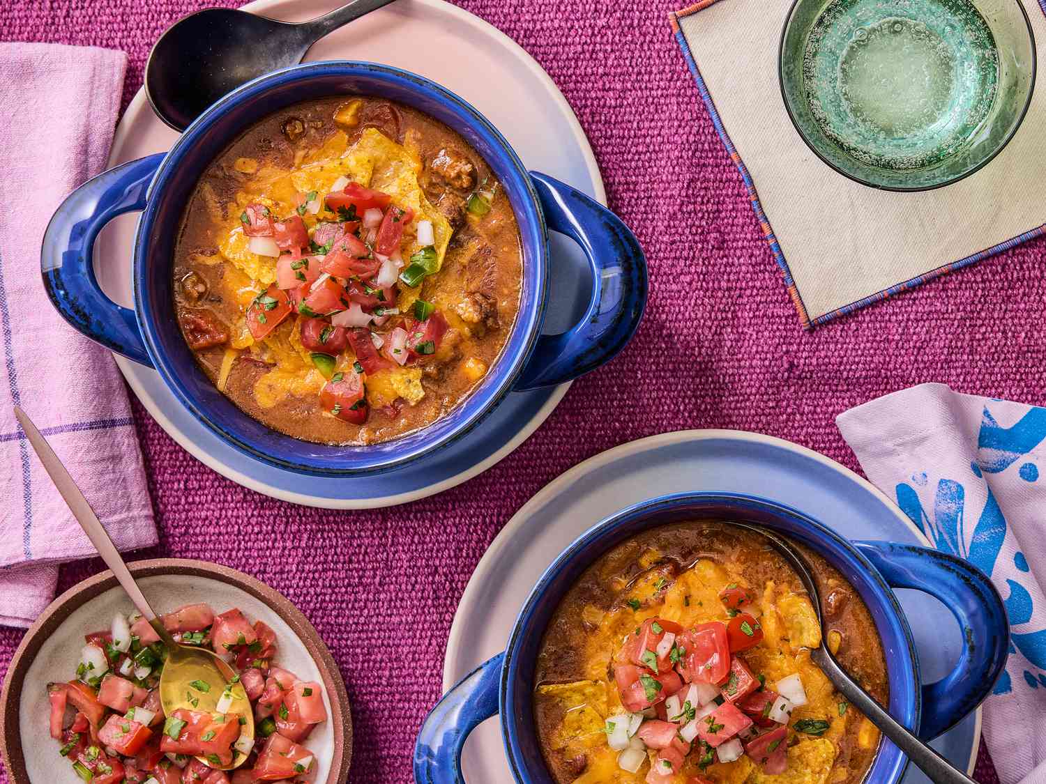 Overhead of 2 blue bowls with taco soup, on with a spoon in it. A small dish with pico de gallo on it. Pink napkins and sparkling water and a magenta colored textile surface. 