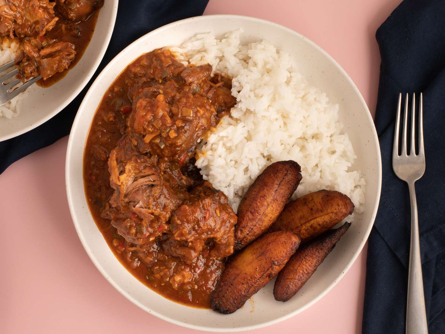 Rabo encendido with white rice and maduros served in a white bowl on a pink countertop.