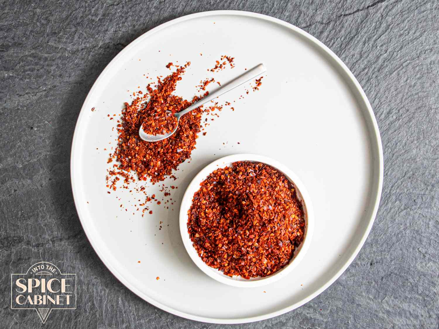 Overhead view of Aleppo Pepper on a plate on a grey background with an Aleppo Pepper logo in the lower left corner