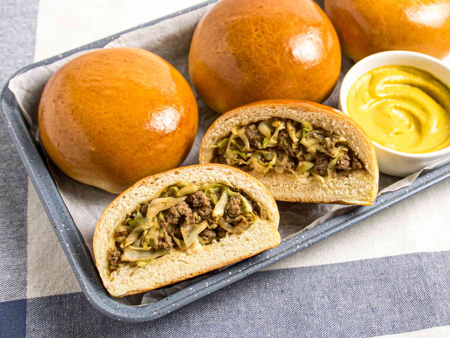 3 Bierocks on a platter with small white mustard dish, with a 4th bun cut open and filling is visible. Platter is on a grey white and blue striped textile. 