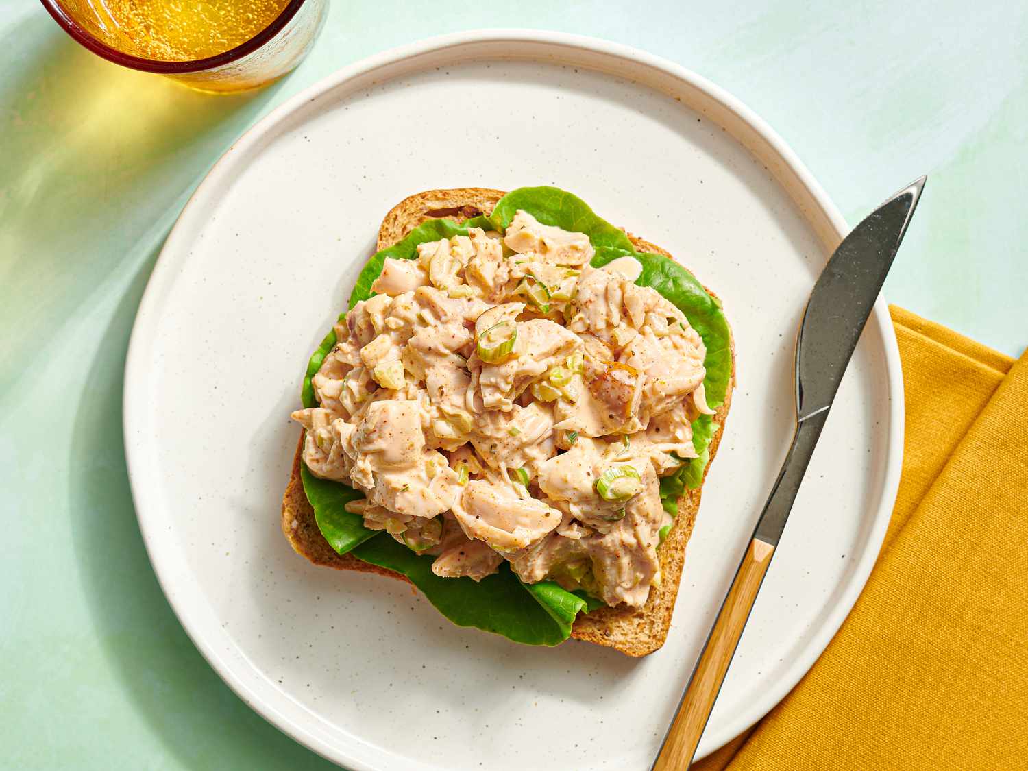 Open-face smoked chicken salad sandwich on a plate with a knife, a glass and orange cloth napkin on the periphery.