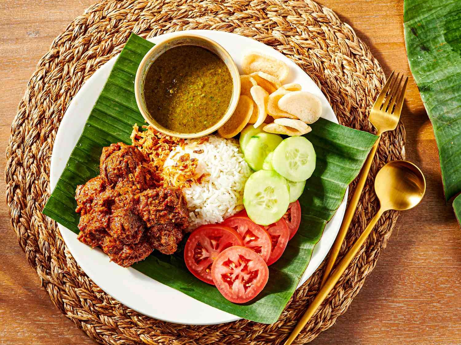 Plate of beef Rendang with rice, cucumbers, tomatoes and crisps on a banana leaf, with a woven placemat and gold cutlery