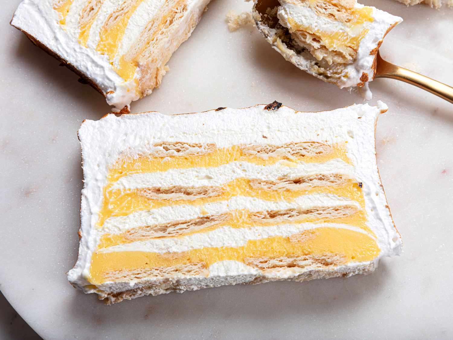 Overhead angle of 2 slices of Lemon Meringue Icebox cake on a marble plate. In the top right corner, there is a spoon with a bite of lemon meringue pie on it 