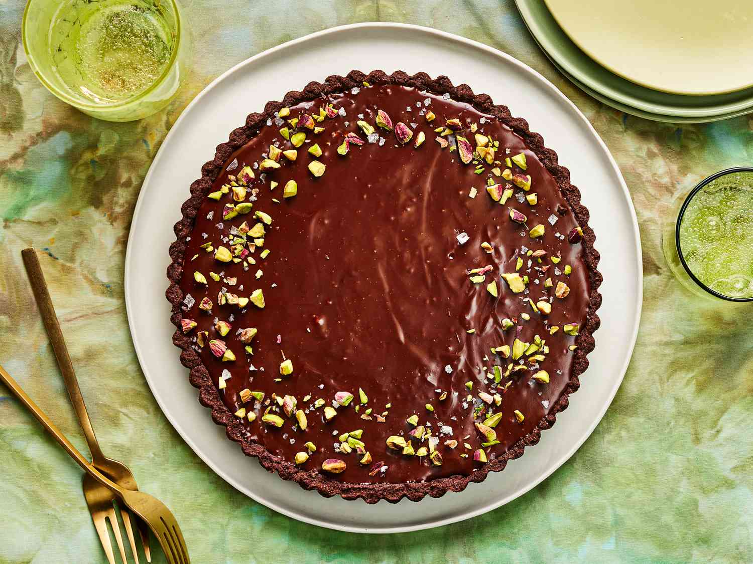 Whole Dubai Chocolate Tart topped with pistachio, on green tablecloth with 2 glasses of water, and stacks of plates in the corner 