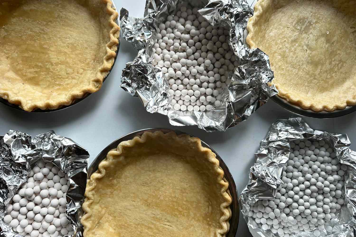 A collection of baked pie crusts and pies filled with ceramic pie weights on a white surface.