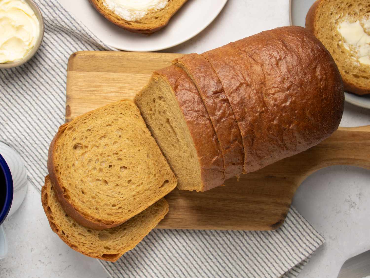 Sliced loaf of anadama bread on a wooden board, with two slices on plates, with grey striped napkin, mug of coffee, plate, butter, and breadknife behind it 