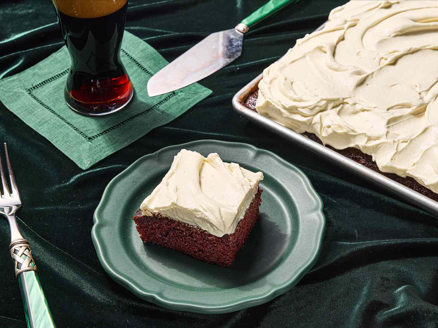 piece of guinnes chocolate cake taken from corner of sheetpan and placed on a green plate. Side of beer on green napkin, green fork, on top of green fabric