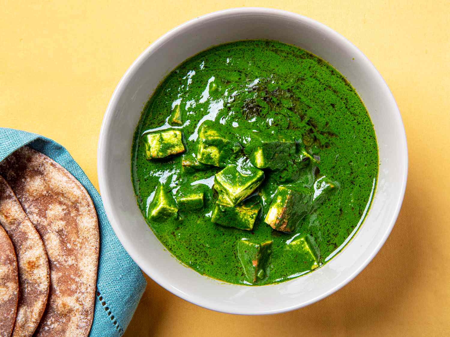 Bright green Palak Paneer in a white bowl one an orange back next to some roti.