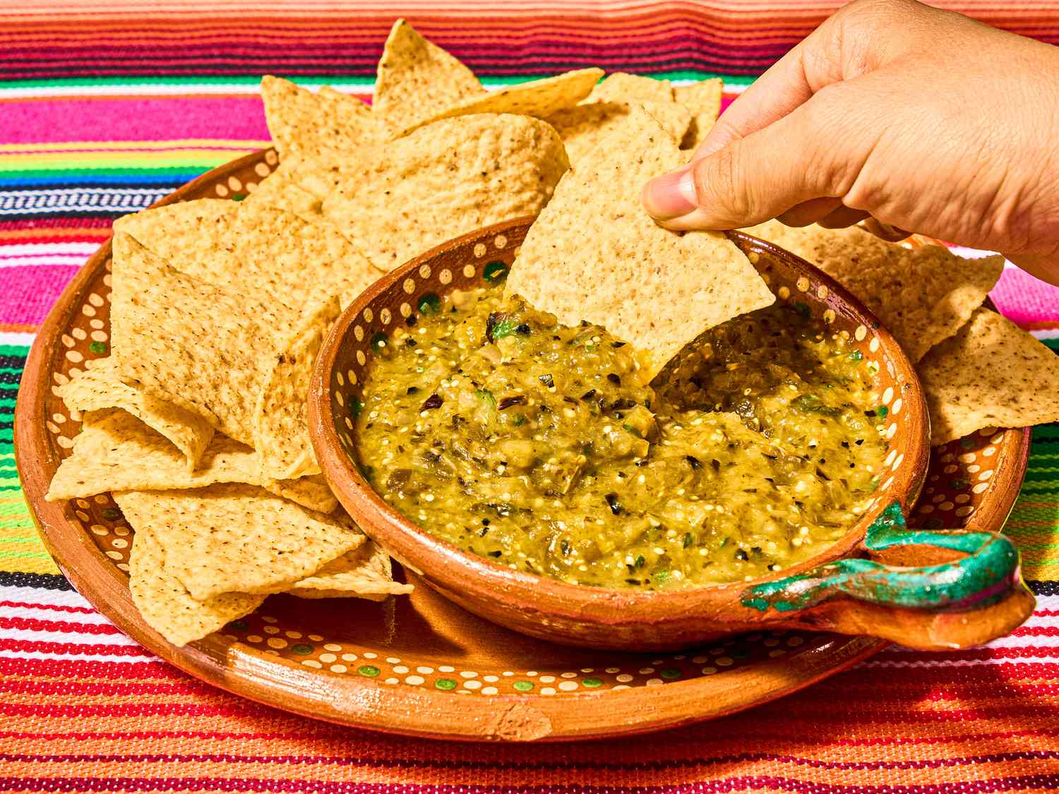 Mexican clay bowls with tortilla chips and charred salsa verde on a colorful traditional Mexican textile, with a hand dipping a chip into the salsa 