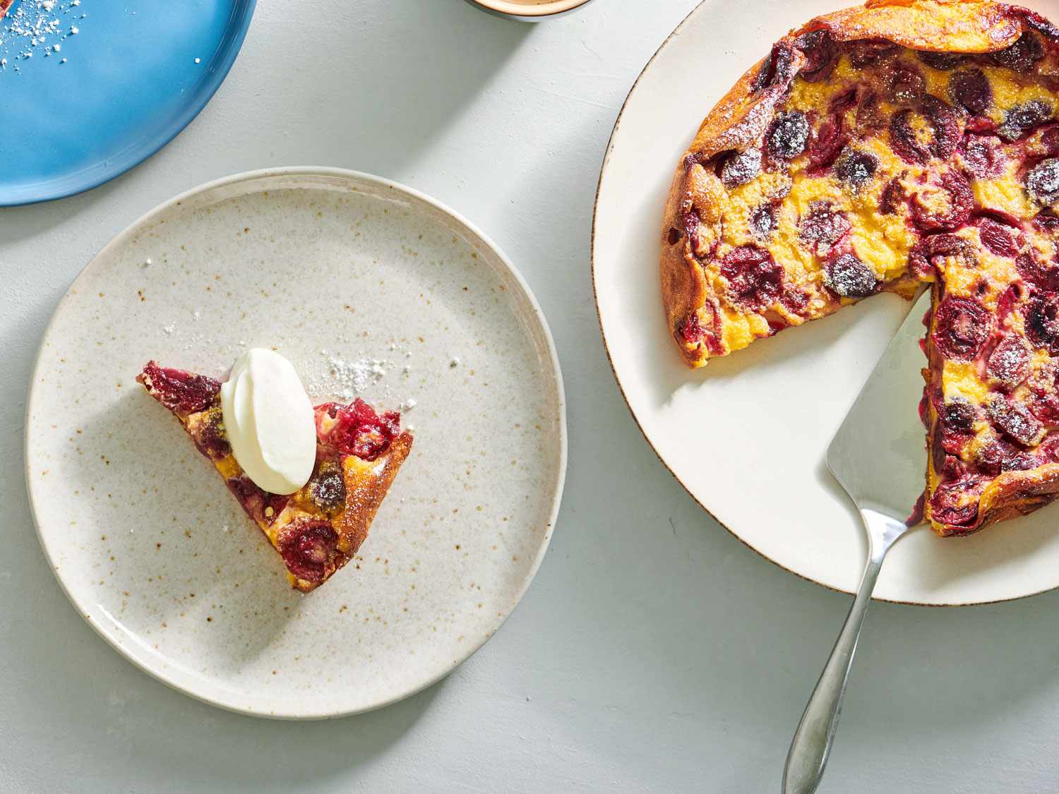 A slice of cherry clafoutis on a speckled ceramic plate on the left side of the image. On the right side is the remaining clafoutis, showing a slice has been removed.