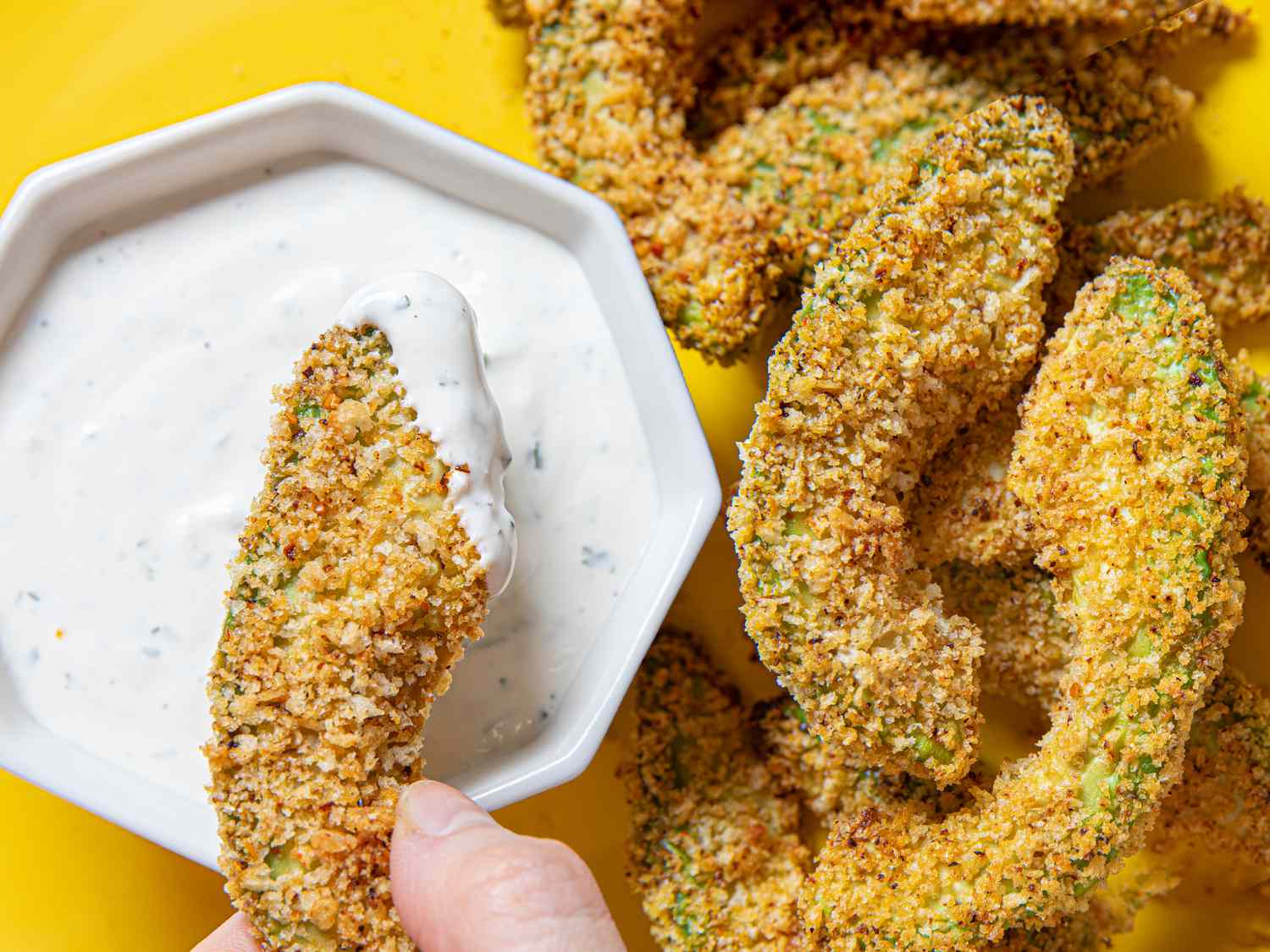 Overhead view of dipping an avocado fry into ranch on a yellow plate