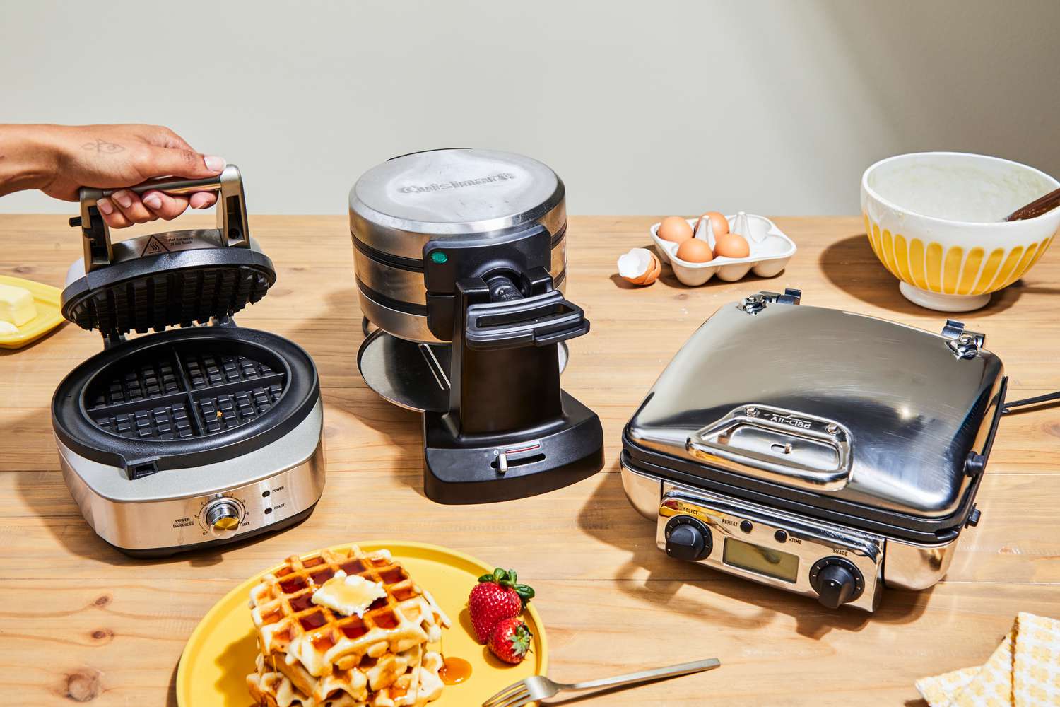 Three waffle makers on a counter, surrounded by breakfast items