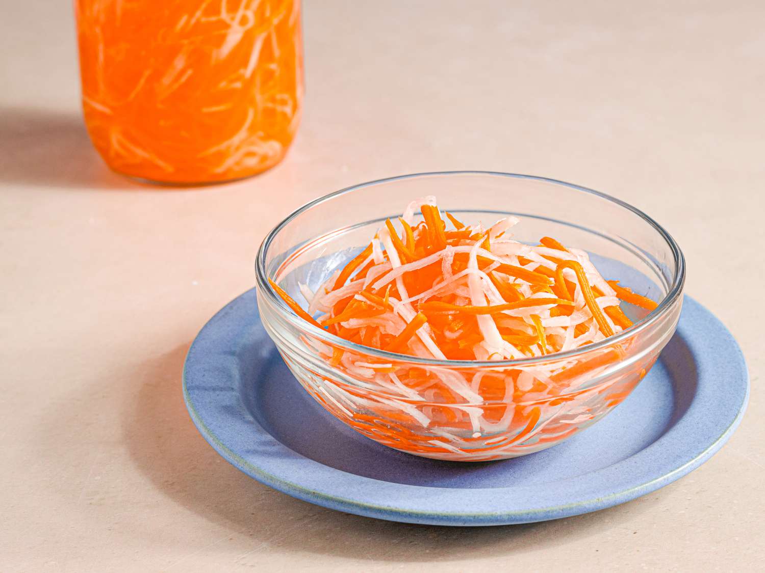 A small glass bowl on a blue plate holding Vietnamese pickled carrots and daikon. There is a jar of pickles and bring in the top lefthand corner of the image.