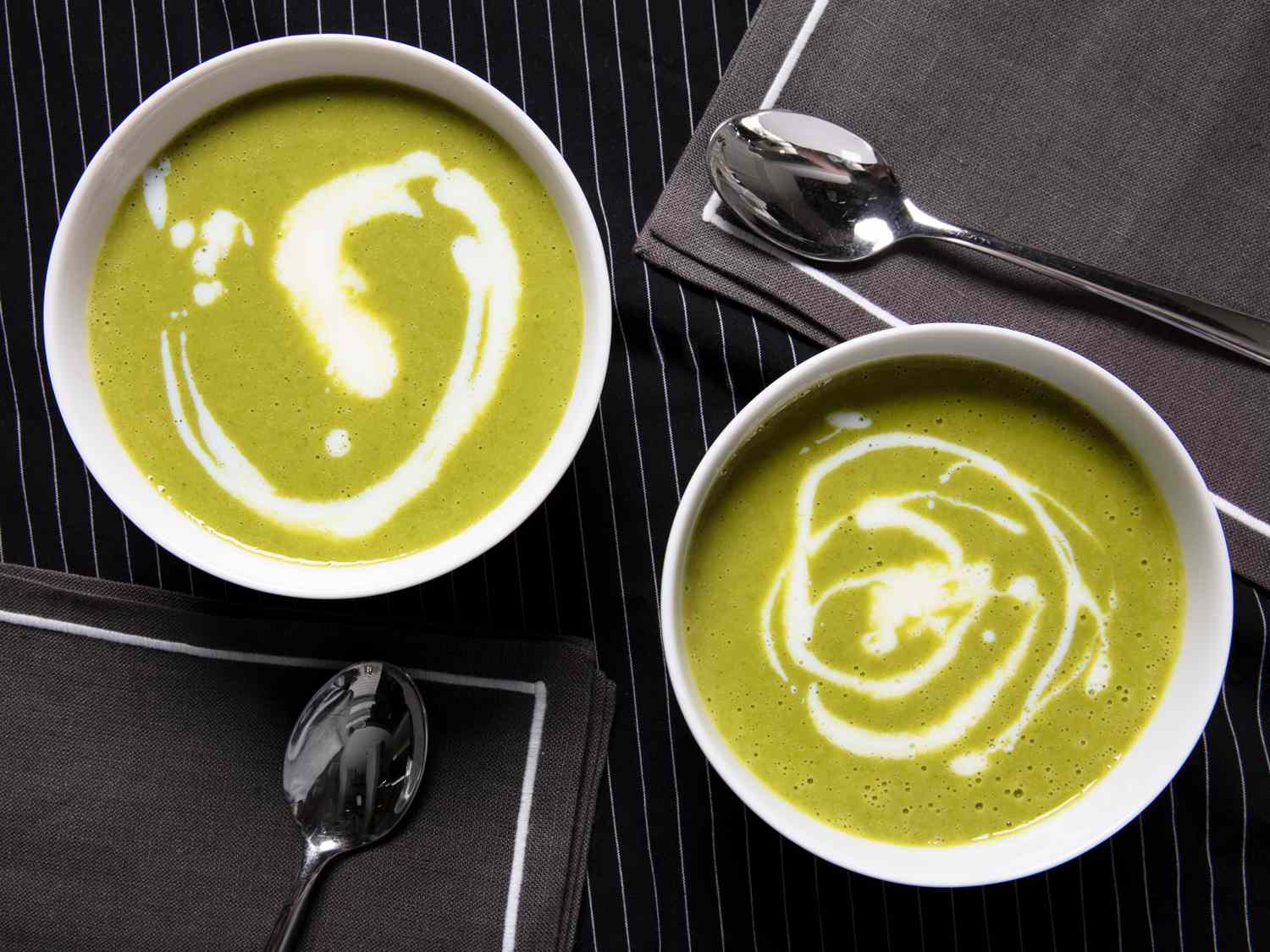 Overhead view of garlic scape soup in two white bowls on a black with thin white stripe background and two grey napkins and spoons