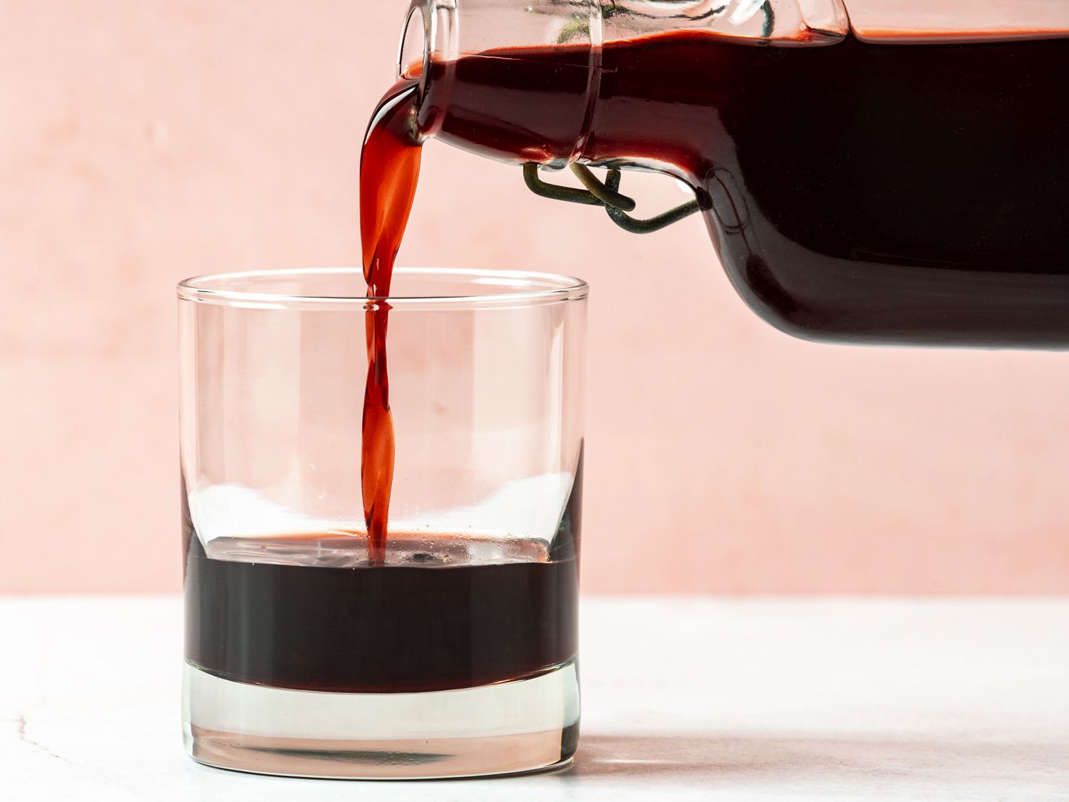 A bottle of DIY cherry liqueur on the right hand side of the image pouring liqueur into a short rocks cocktail glass on the left hand side of the image.