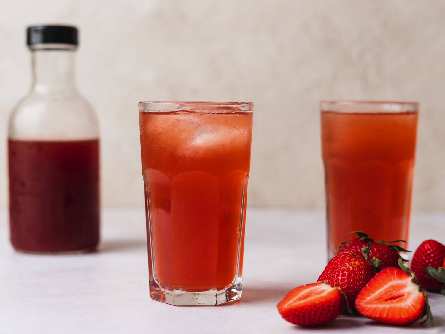 Two glasses of berry shrub. On the right hand side of the image are some strawberries, both whole and sliced in half, and on the left side of the image, in the background, is a small glass bottle holding more shrub.