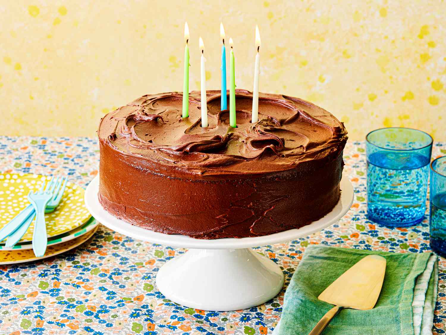 A chocolate-frosted layer cake with billowing swirls of frosting, birthday candles, accompanied by serving plates, forks, and blue glasses of water against a yellow background.