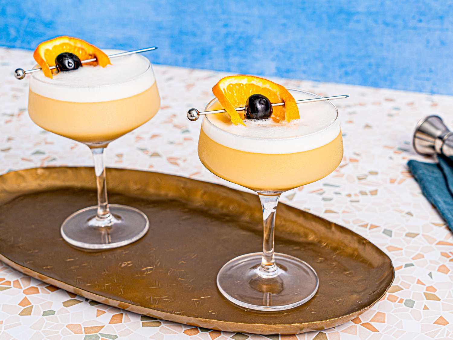 Two whiskey sour cocktails, garnished with an orange wheel and a cherry on a cocktail pick, served on a tray